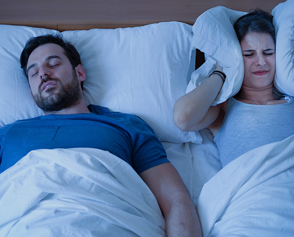 Couple in bed; man snoring while woman covers ears with pillow, illustrating sleep disruption issues.