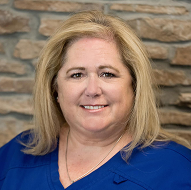 Professional headshot of woman with blonde hair smiling at camera against brick wall background.