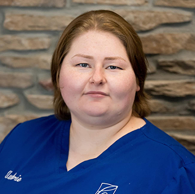 Woman in blue dental scrubs smiling at camera against brick wall background.