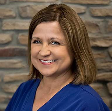 Professional headshot of woman in blue scrubs smiling at camera against brick wall background.
