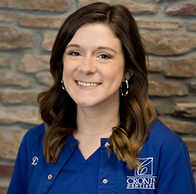 Smiling female dental professional in blue Cronin Dentistry uniform against brick background.