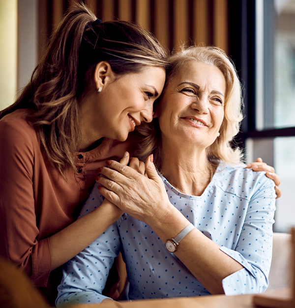 Adult daughter hugging smiling senior mother from behind in bright home setting.