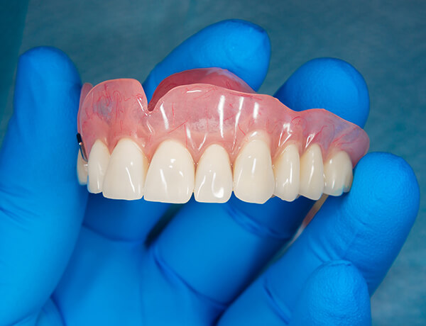 Dental professional holding upper denture with artificial teeth and pink gum base against blue gloved hand.