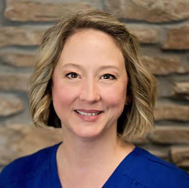 Woman smiling at camera wearing blue shirt against brick wall background.