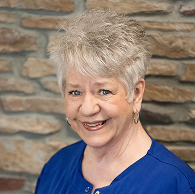 Professional headshot of smiling woman with gray hair in blue top against brick wall background.