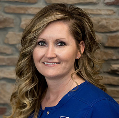 Professional headshot of woman smiling, wearing blue scrubs, brick wall background.