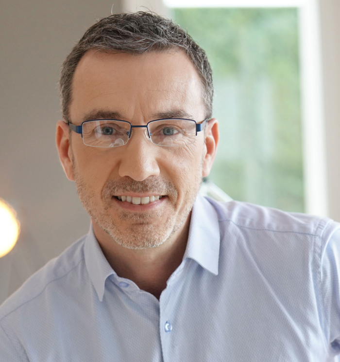 Professional headshot of smiling man wearing glasses and light blue shirt in dental office setting.