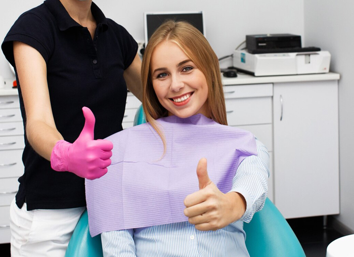 Dental patient giving thumbs up while dentist in pink gloves stands behind in modern office setting.
